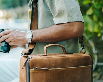 May include: A brown leather messenger bag with a top handle and a long adjustable strap. The bag has a zippered closure and a front pocket.
