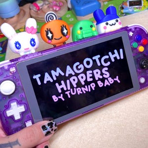 May include: A purple Nintendo Switch Lite console with a clear case and colorful character charms attached. The screen displays the text "TAMAGOTCHI HIP/PERS BY TURNIP BABY".
