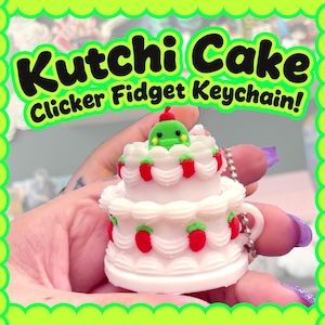 Può includere: Un portachiavi fidget clicker a forma di torta bianca e rossa con una creatura verde sopra. Il portachiavi ha una catena d'argento e il testo "Kutchi Cake Clicker Fidget Keychain!" in verde e nero.