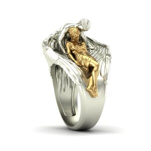 K&ouml;nnte beinhalten: Ein silberner Ring mit einer goldfarbenen Figur, die auf silbernen Fl&uuml;geln liegt, mit einer weiteren silbernen Figur dar&uuml;ber. Der Ring hat ein breites Band und ein detailliertes, geformtes Design. Der Ring ist vor wei&szlig;em Hintergrund.