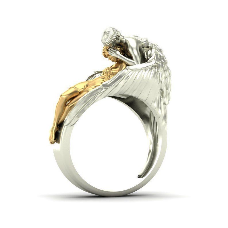 K&ouml;nnte beinhalten: Ein silberner und goldener Ring mit einem Engel, der eine Figur h&auml;lt. Der Ring hat ein detailliertes Design mit den Fl&uuml;geln des Engels und der Form der Figur. Der Ring ist vor einem wei&szlig;en Hintergrund.