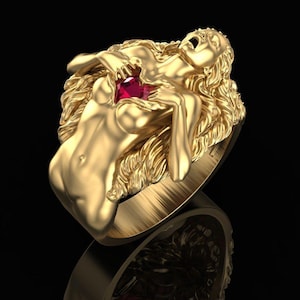 Peut inclure: Une bague dorée représentant une figure féminine sculptée avec des cheveux fluides, tenant une pierre précieuse rouge. La bague présente des détails complexes et une finition polie, sur fond sombre.