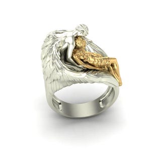 K&ouml;nnte beinhalten: Ein silberner Ring mit einem skulpturalen Design von zwei Figuren, eine silberne und eine goldene, die mit einem Fl&uuml;gelmotiv verflochten sind. Der Ring hat ein breites Band und ein detailliertes, k&uuml;nstlerisches Aussehen.