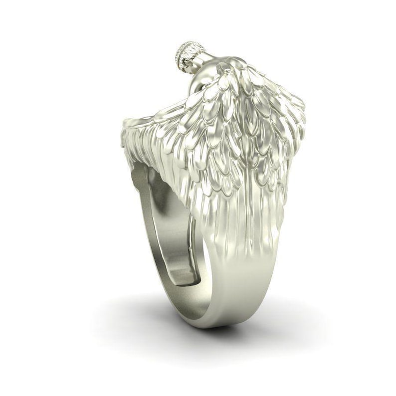 K&ouml;nnte beinhalten: Ein silberfarbener Ring mit einem Engel mit ausgebreiteten Fl&uuml;geln. Der Ring hat ein detailliertes Design, wobei die Fl&uuml;gel und der K&ouml;rper des Engels aufwendig gestaltet sind. Der Ring wird vor einem wei&szlig;en Hintergrund gezeigt, um seinen metallischen Glanz hervorzuheben.