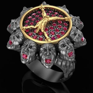 May include: A detailed ring featuring a gold figure in a crucifixion pose within a gold-rimmed oval, surrounded by red gems. The ring's band is adorned with multiple screaming faces in a dark metallic finish, creating a dramatic and artistic statement.