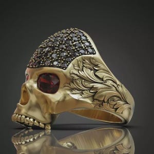 May include: A gold-toned ring shaped like a human skull. The top of the skull is encrusted with numerous small black stones, while the eye sockets feature deep red stones. The side of the ring band is decorated with intricate, raised leaf-like carvings.