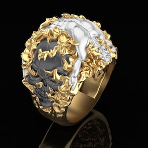 May include: A gold-toned ring featuring a detailed design. The ring showcases a black skull with a white figure on top, surrounded by gold-colored flames. The ring's band is also gold-toned, creating a striking contrast against the dark background.