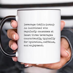 May include: White ceramic mug with a black handle and black rim. The mug has a definition of a "Beverage Goblin" printed on it in black text. The definition reads: "An individual who regularly consumes at least three beverages concurrently, typically for hydration, caffeine, and enjoyment."