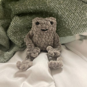 May include: A gray crocheted frog plush toy sitting on a white surface with a green blanket behind it.