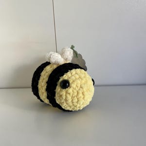 May include: A small, round, handmade crochet bee plush toy featuring alternating horizontal stripes of soft yellow and black yarn. The bee has two small white textured wings on its back and two round, black plastic safety eyes on its face.