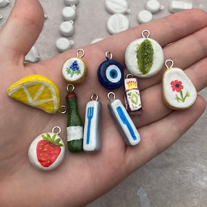 May include: A collection of handmade charms in various shapes and designs. Includes a yellow lemon slice, a red strawberry, a blue evil eye, a green bottle, and a white charm with a red flower. Each charm has a silver loop for attaching to jewelry.