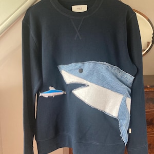 May include: Navy blue sweatshirt featuring a shark design. The shark is crafted from denim and white fabric, with a smaller fish applique. The label displays the text "M&S".