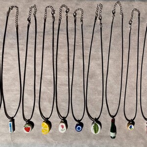May include: A collection of pendant necklaces with black cords and silver-tone chains. The pendants feature various designs, including a fork, knife, strawberry, lemon, flower, evil eye, leaf, wine bottle, and dice.