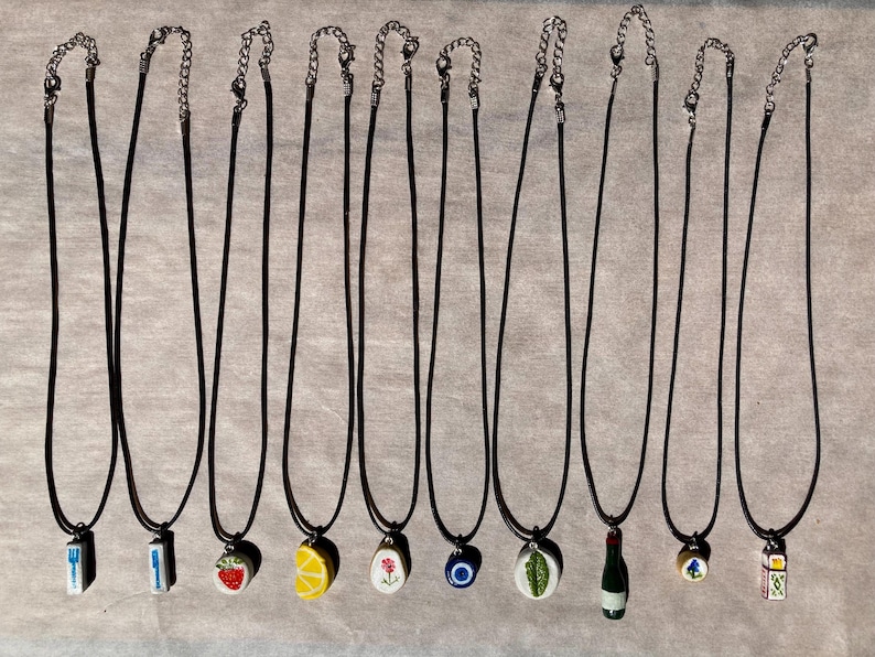 May include: A collection of pendant necklaces with black cords and silver-tone chains. The pendants feature various designs, including a fork, knife, strawberry, lemon, flower, evil eye, leaf, wine bottle, and dice.