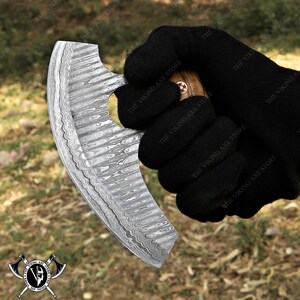 May include: A curved, stainless steel knife with a wooden handle. The blade has a hammered finish and a small hole near the handle. The knife is held in a gloved hand.