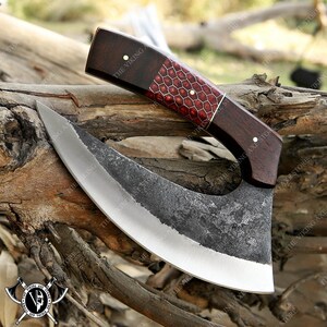 May include: A curved, hand-forged knife with a dark gray blade and a wooden handle. The handle is decorated with a red and gold honeycomb pattern. The knife is resting on a piece of wood.