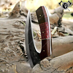 May include: A curved black axe with a wooden handle. The handle is decorated with a red and gold pattern. The axe is resting on a wooden surface.