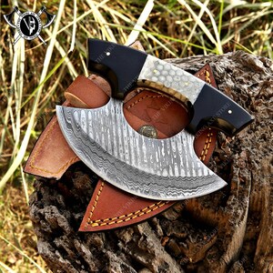 May include: A Damascus steel cleaver with a black and white handle. The cleaver is in a brown leather sheath with stitching. The cleaver is on a wooden surface.