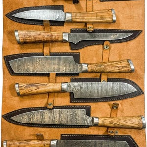 Può includere: Un set di sette coltelli da cucina con lame in acciaio damasco e manici in legno chiaro venato, ciascuno riposto in un fodero in pelle marrone scuro fissato su un grande tappetino avvolgibile in pelle color cuoio.