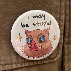 May include: A round white button with the text "i may be stupid" in black. Below the text is a cartoon illustration of a boar's head with blue eyes, a pink snout, and tusks. Two yellow stars flank the boar's head.
