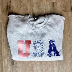 May include: A heather gray sweatshirt with the letters "USA" appliqued on the front. The "U" is red gingham, the "S" is blue floral, and the "A" is blue floral. The sweatshirt is folded on a wooden surface.