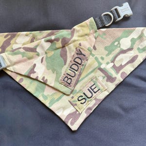 May include: Camouflage dog bandana with the names "Buddy" and "Sue" embroidered on patches. The bandana is paired with a black collar with a gray buckle. The fabric features a mix of green, brown, and tan colors.