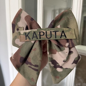 May include: A camouflage fabric bow with the name "KAPUTA" embroidered in black. The bow features a mix of green, brown, and tan colors, and is likely a hair accessory or decorative item.