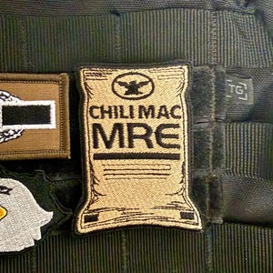 May include: Tan and black embroidered patch with the text "CHILI MAC MRE" and an eagle emblem. The patch is rectangular with a curved bottom edge and is attached to a black tactical vest.