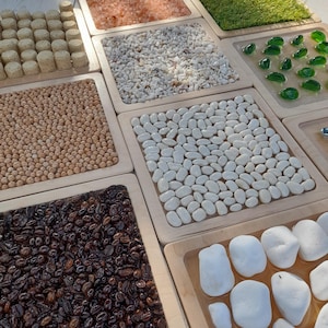 Könnte beinhalten: Ein Set aus neun sensorischen Tabletts mit verschiedenen Texturen, darunter Kaffeebohnen, Kichererbsen, weiße Bohnen, kleine weiße Steine, rosa Himalaya-Salz, weiße Kieselsteine, grünes Gras und Glasperlen. Die Tabletts sind aus Holz gefertigt und haben eine hellbraune Oberfläche.