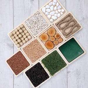 May include: Nine wooden trays with different textures and materials, including white stones, brown rope, wood rounds, chickpeas, buckwheat, black beans, green grass, and a green surface.