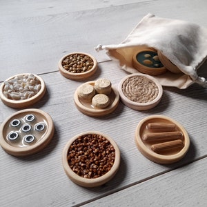 Könnte beinhalten: Ein Satz von acht Holz-Sinnestabletts mit verschiedenen Texturen und Materialien. Die Tabletts sind gefüllt mit Gegenständen wie Linsen, Buchweizen, kleinen Holzperlen und kleinen Holzstiften. Die Tabletts sind auf einer hellen Holzoberfläche angeordnet.