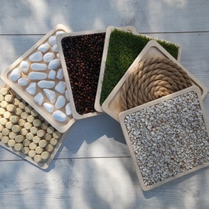 May include: Six wooden trays with different textures, including white stones, coffee beans, green grass, rope, small white stones, and cork rounds.