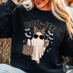 Puede incluir: Una sudadera negra de manga larga con el gr&aacute;fico de un fantasma con lazo y gafas de sol de estampado de leopardo, sosteniendo un bolso y un vaso de caf&eacute;. El texto BOO-JEE aparece impreso arriba, rodeado de peque&ntilde;os murci&eacute;lagos y estrellas.