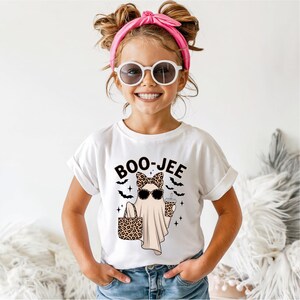 Puede incluir: Una camiseta blanca de manga corta con el gr&aacute;fico de un fantasma con lazo y gafas de sol de estampado de leopardo, que sostiene un bolso y un vaso a juego. El texto BOO-JEE est&aacute; impreso encima del fantasma junto a peque&ntilde;os murci&eacute;lagos.