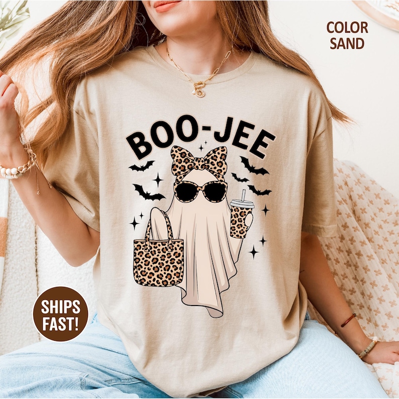 Puede incluir: Una camiseta color arena con el gr&aacute;fico de un fantasma que lleva un lazo y gafas de sol con estampado de leopardo, sosteniendo un bolso y un vaso a juego. El texto BOO-JEE aparece arriba, rodeado de murci&eacute;lagos negros y estrellas.