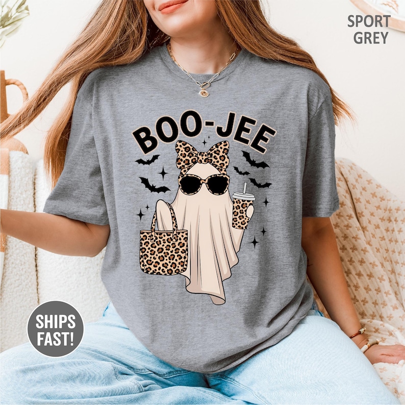 Puede incluir: Una camiseta gris sport con el gr&aacute;fico de un fantasma con lazo y gafas de sol de estampado de leopardo, sosteniendo un bolso y un vaso a juego. El texto BOO-JEE aparece impreso encima, rodeado de peque&ntilde;os murci&eacute;lagos negros y estrellas.