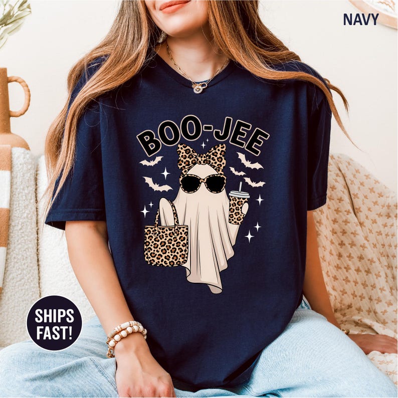 Puede incluir: Una camiseta azul marino con el gr&aacute;fico de un fantasma que lleva un lazo y gafas de sol, sosteniendo un bolso y un vaso de caf&eacute; con estampado de leopardo. El texto BOO-JEE aparece sobre el fantasma, rodeado de murci&eacute;lagos y estrellas.
