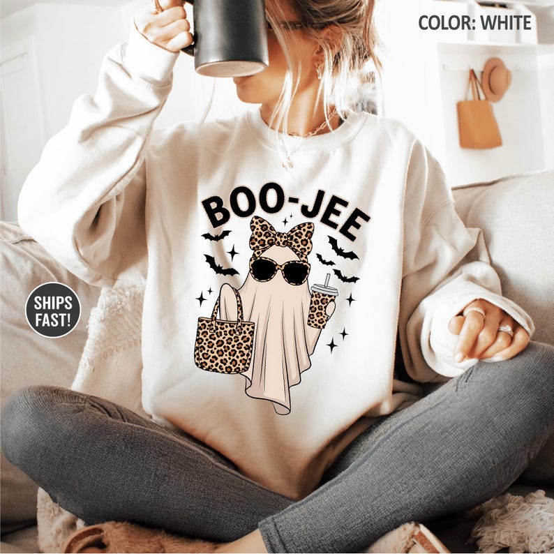 Puede incluir: Una sudadera blanca de cuello redondo con un gr&aacute;fico de un fantasma con lazo y gafas de sol de estampado de leopardo, sosteniendo un bolso y un vaso de caf&eacute;. El texto BOO-JEE aparece impreso encima, rodeado de murci&eacute;lagos y estrellas negras.