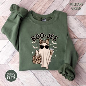 Puede incluir: Una sudadera de cuello redondo color verde militar con un gr&aacute;fico de un fantasma con gafas de sol, un lazo de estampado de leopardo, y sosteniendo un bolso y una taza de leopardo. El texto BOO-JEE est&aacute; impreso arriba, rodeado de murci&eacute;lagos.