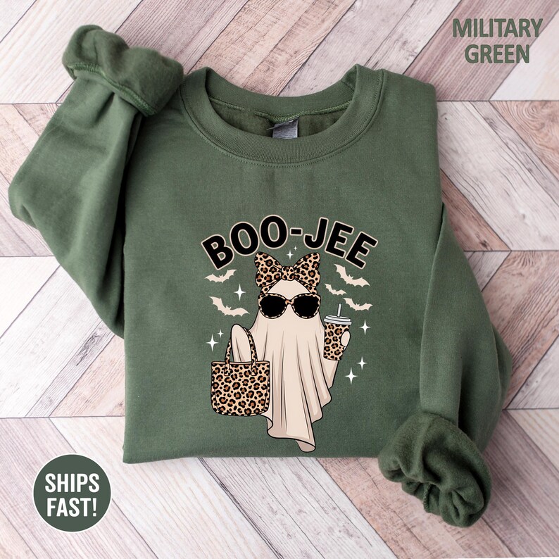 Puede incluir: Una sudadera de cuello redondo color verde militar con un gr&aacute;fico de un fantasma con gafas de sol, un lazo de estampado de leopardo, y sosteniendo un bolso y una taza de leopardo. El texto BOO-JEE est&aacute; impreso arriba, rodeado de murci&eacute;lagos.