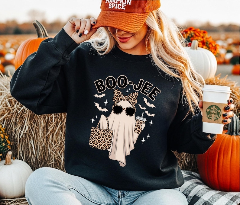 Puede incluir: Una sudadera negra de manga larga con el gr&aacute;fico de un fantasma con lazo y gafas de sol de estampado de leopardo, sosteniendo un bolso y un vaso de caf&eacute;. El texto BOO-JEE aparece impreso arriba, rodeado de peque&ntilde;os murci&eacute;lagos y estrellas.