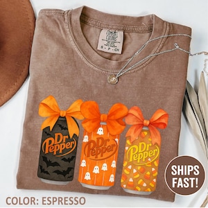 May include: An espresso-colored t-shirt featuring a graphic of three soda cans, each topped with an orange bow. The cans display Dr Pepper branding and patterns of black bats, white ghosts, and orange candy corn. The shirt has a Comfort Colors label.