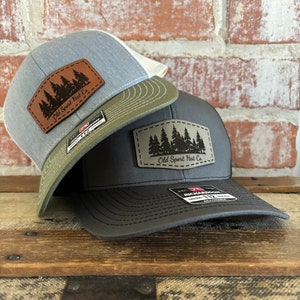 May include: Two baseball caps with a forest silhouette patch that reads "Old Sport Hat Co.". The top cap is light blue with a white mesh back and a brown leather patch. The bottom cap is black with a green mesh back and a brown leather patch.