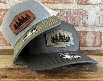 May include: Two baseball caps with a forest silhouette patch that reads "Old Sport Hat Co.". The top cap is light blue with a white mesh back and a brown leather patch. The bottom cap is black with a green mesh back and a brown leather patch.