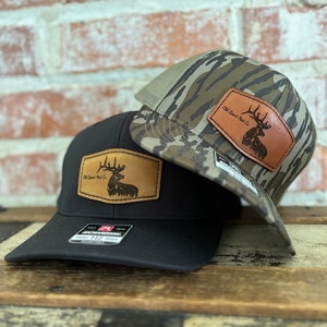 Puede incluir: Dos gorras de camionero con estampado de camuflaje y una gorra de tela negra. Las gorras tienen un parche de cuero con una silueta de ciervo y el texto "Old Sport Hat Co."