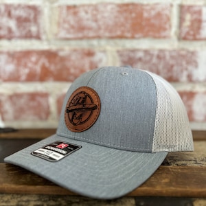 Op de afbeelding: Een grijze en witte truckercap met een bruine leren patch met een gestileerd ontwerp van een sigaar en een schedel. De pet heeft een mesh achterkant en een gebogen klep.