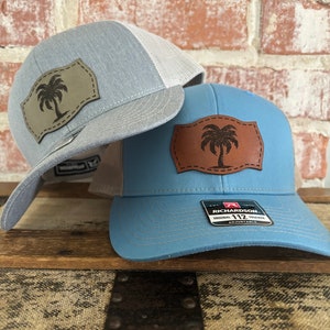 May include: Two trucker hats with a palm tree patch. The hat on the left is light blue with a white mesh back. The hat on the right is blue with a white mesh back. Both hats have a brown leather patch with a palm tree design. The hat on the right has a black label that says "Richardson 112 Original Trucker Adjustable".