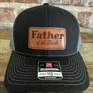 Peut inclure: Casquette de camionneur noire et grise avec un patch en cuir marron qui dit "Father of the Bride". La casquette est réglable et porte le logo Richardson 112.