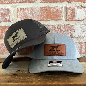 Puede incluir: Dos gorras de camionero con un parche de cuero marrón que dice "Old Sport Hat Co." y una silueta de un perro. Una gorra es negra con una malla negra en la parte posterior y la otra es gris con una malla gris en la parte posterior. La gorra gris tiene una etiqueta negra que dice "Richardson 112 Original Trucker Adjustable".