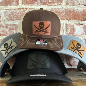 Puede incluir: Tres gorras de camionero con malla marrón, negra y gris en la parte posterior. Cada gorra tiene un parche de cuero marrón con un diseño de calavera y huesos cruzados. Las gorras están apiladas una encima de la otra.